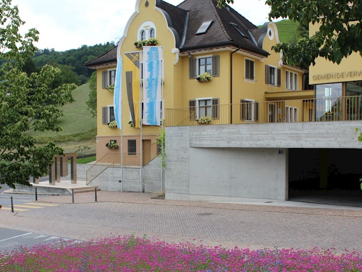 Blau-weisse Energiestadtfahnen vor dem Gemeindehaus Mauren. Im Vordergrund blühen Blumen.