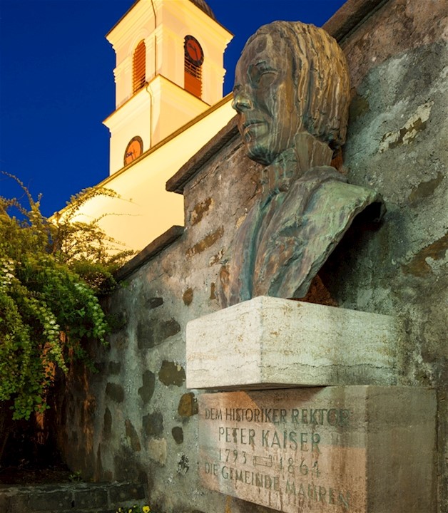 Bronzebüste von Peter Kaiser an der Kirchenmauer. Im Hintergrund der beleuchtete Kirchturm von St. Peter und Paul